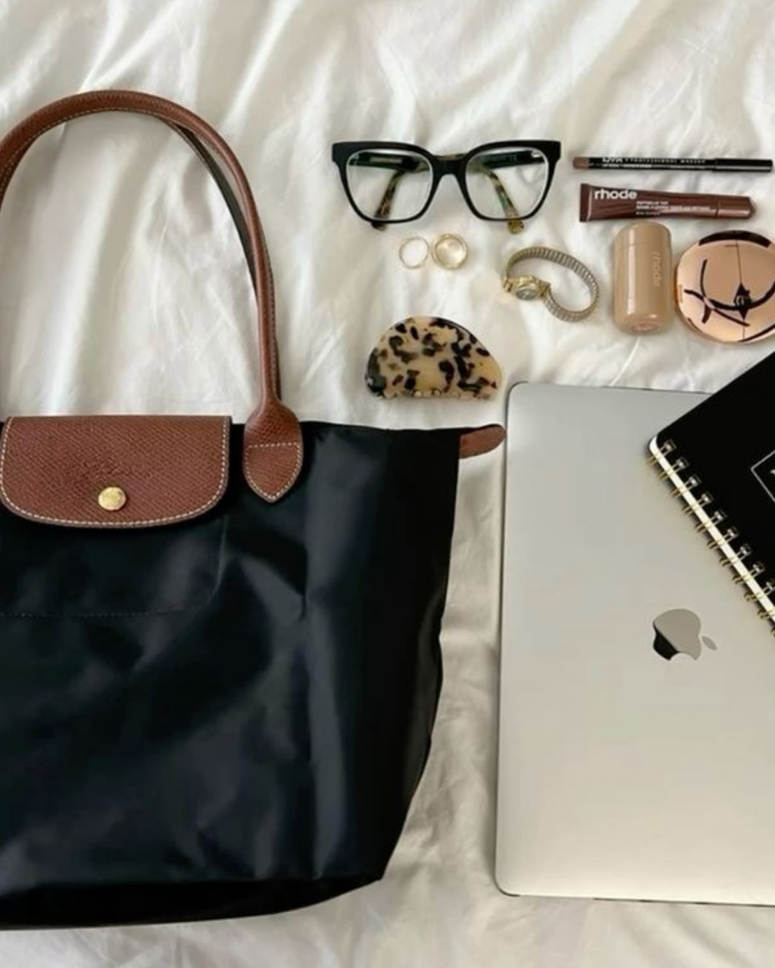 Black handbag with accessories including glasses, makeup items, and a laptop on a white surface.