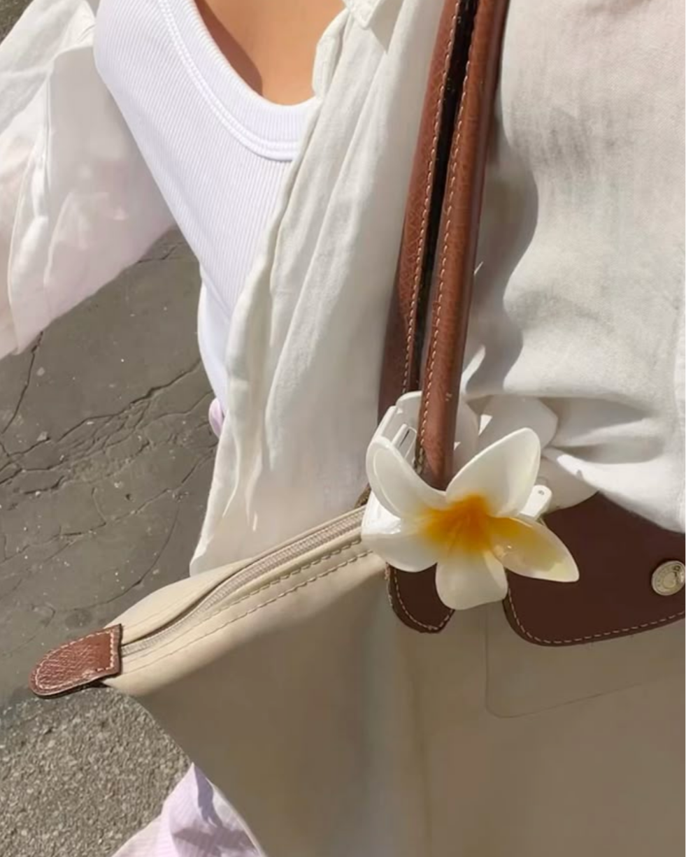 Beige handbag with a white flower accessory held by a person wearing a white shirt.